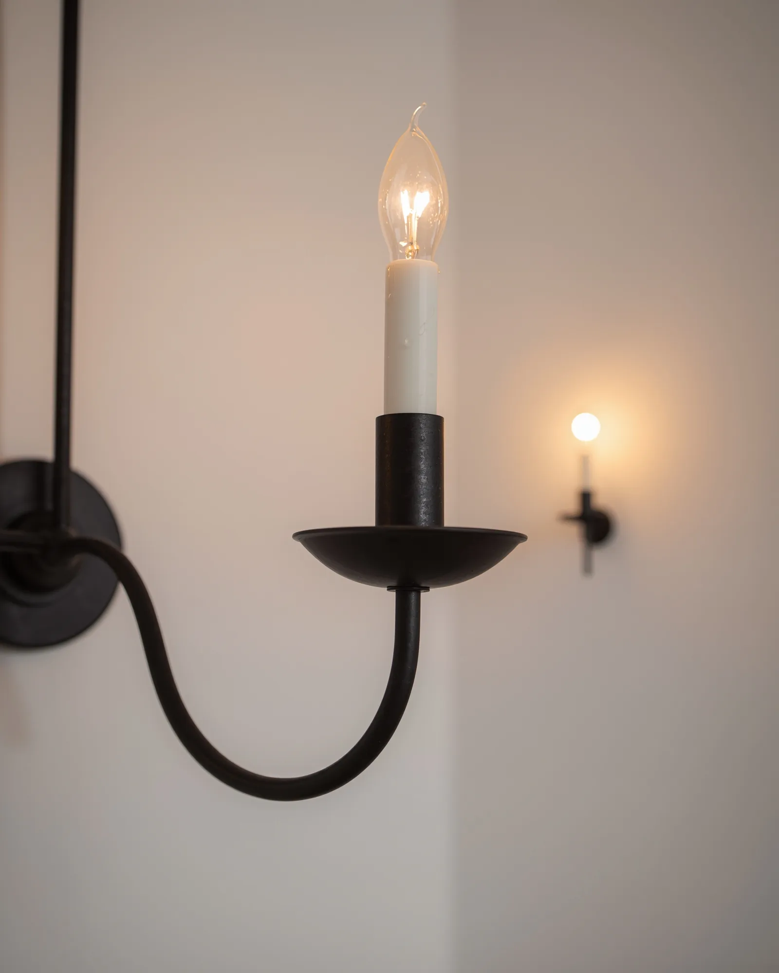 Curved black iron chandelier arm holding a faux candle sleeve and glowing flame-tip bulb, whitewashed room soft behind